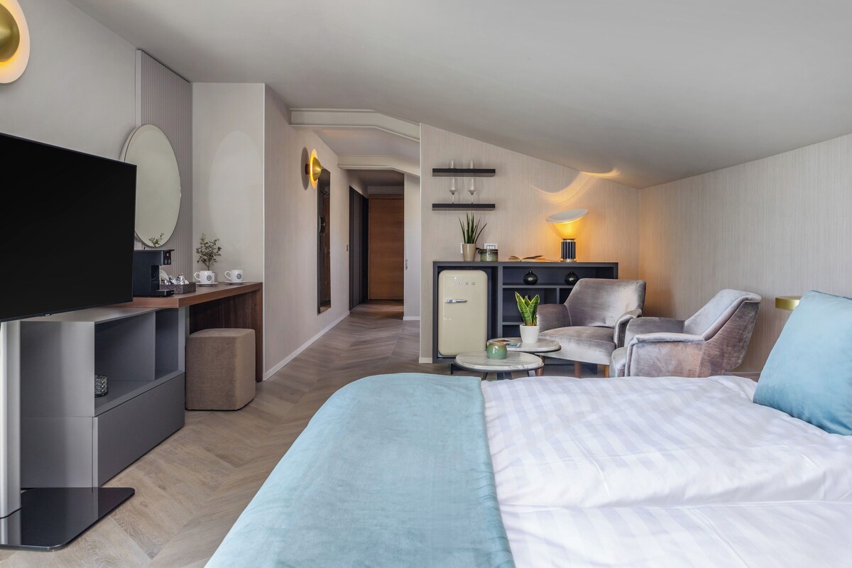A modern hotel room with a large bed, a desk with a flat-screen TV, a seating area with chairs, and a small kitchenette.