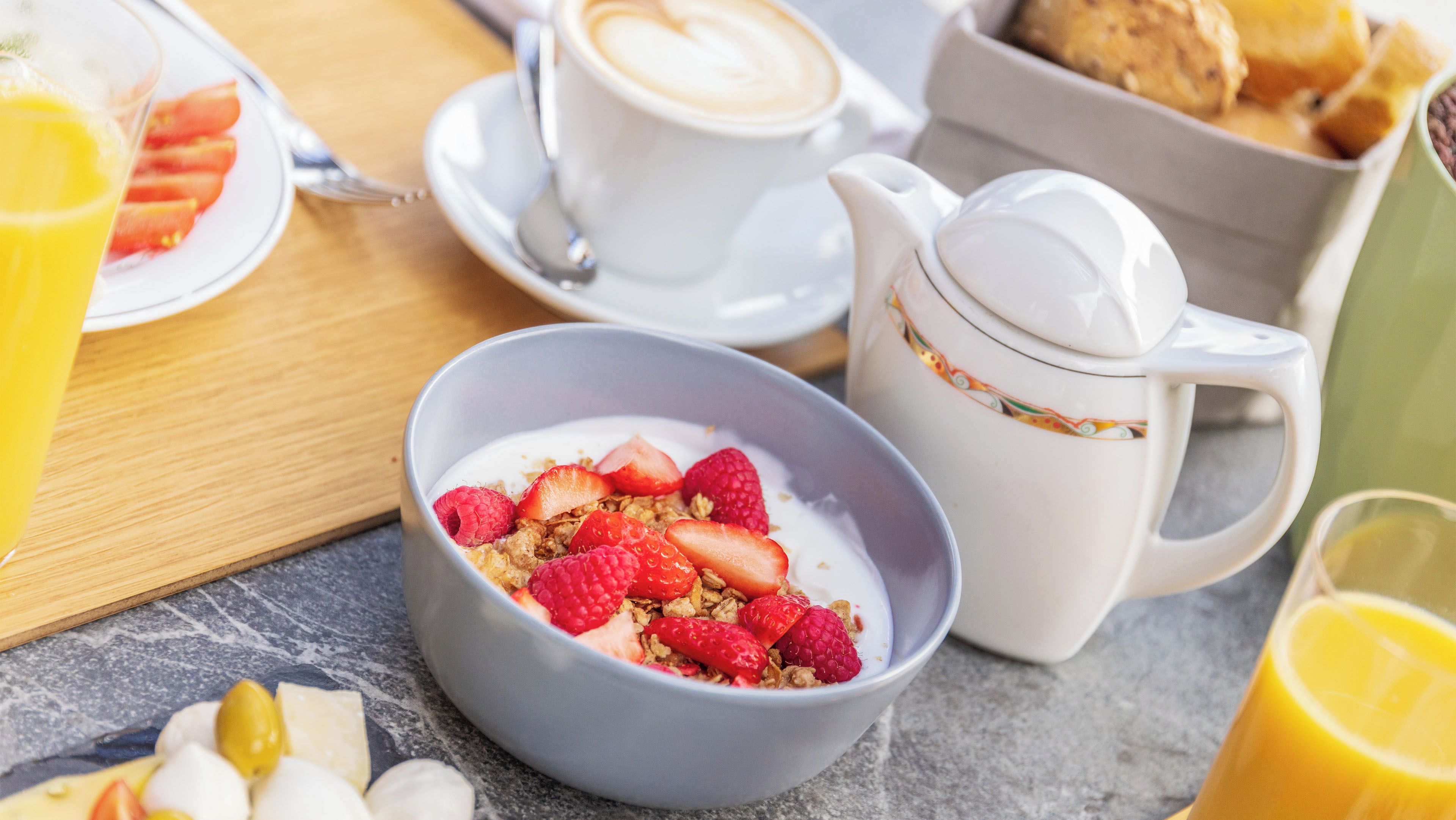 Un desayuno con un tazón de yogur cubierto de fresas y granola, una taza de café y una tetera sobre una mesa.