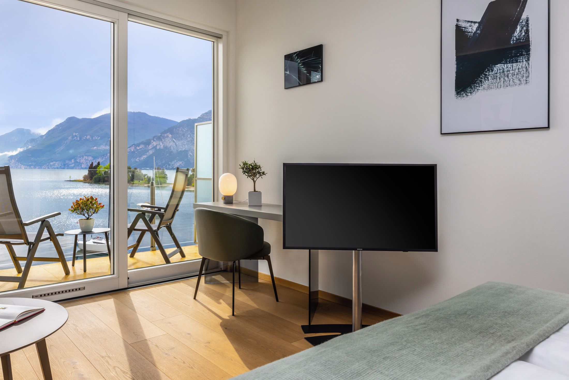 A modern hotel room with a large window offering a scenic mountain view, a flat-screen TV, a desk with a chair, and a bed with a green blanket.