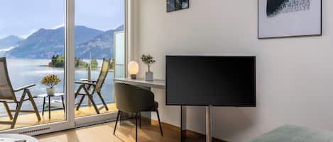 A modern hotel room with a large window offering a scenic mountain view, a flat-screen TV, a desk with a chair, and a bed with a green blanket.