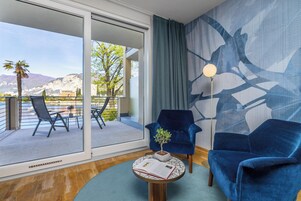 Una habitación moderna con un amplio ventanal corredero que da a un balcón con mobiliario exterior y vistas a montañas.