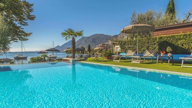 A clear blue pool with lounge chairs and umbrellas, a view of the sea, and mountains in the background. - Hotel Val di Sogno (Malcesine)