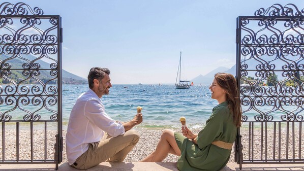 A couple sitting on a pebbled beach with a view of the sea and boats. - Hotel Val di Sogno (Malcesine)