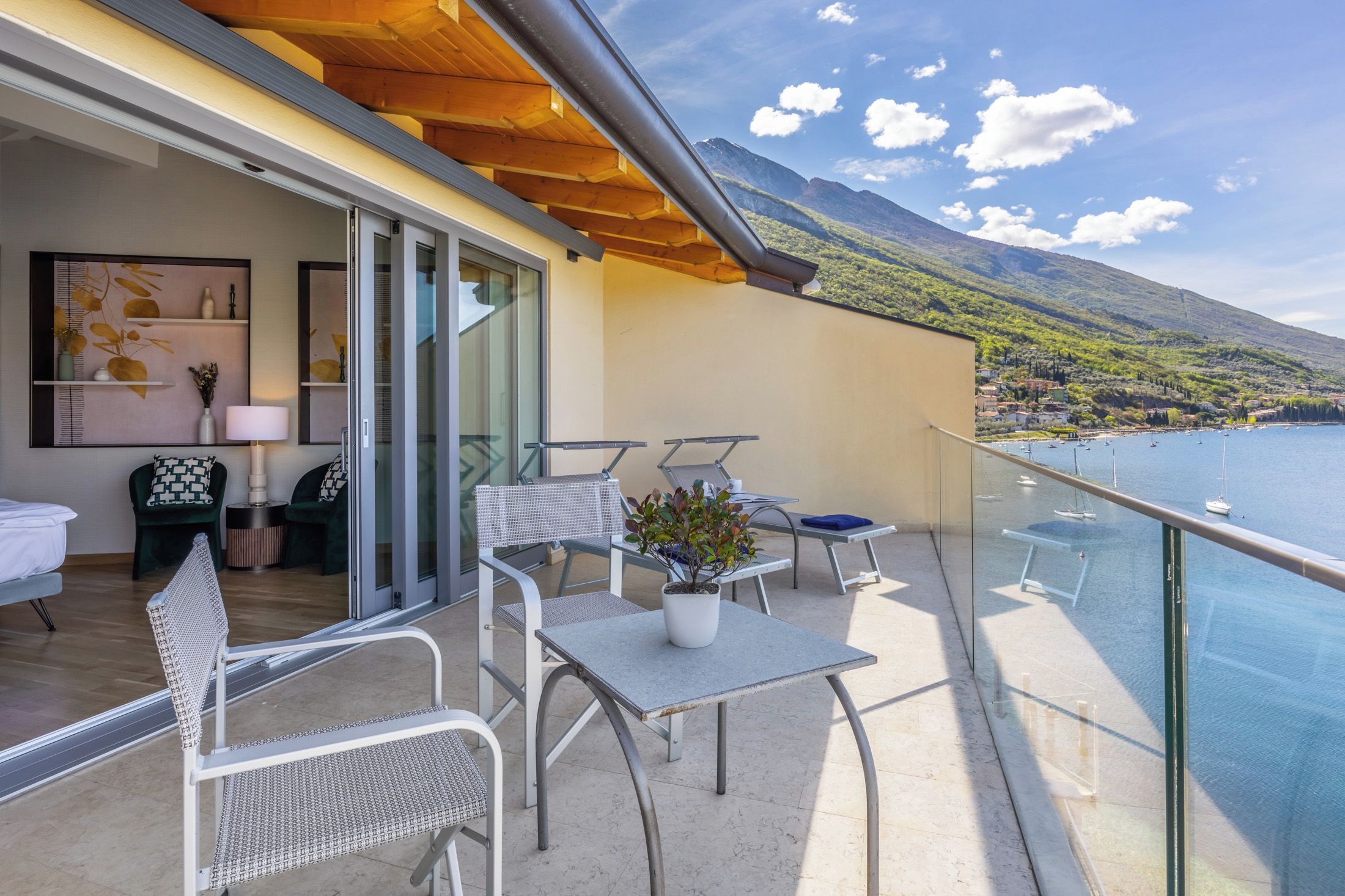 Ein Balkon mit Tisch und Stühlen, einem Blumentopf und Blick auf einen See und Berge.