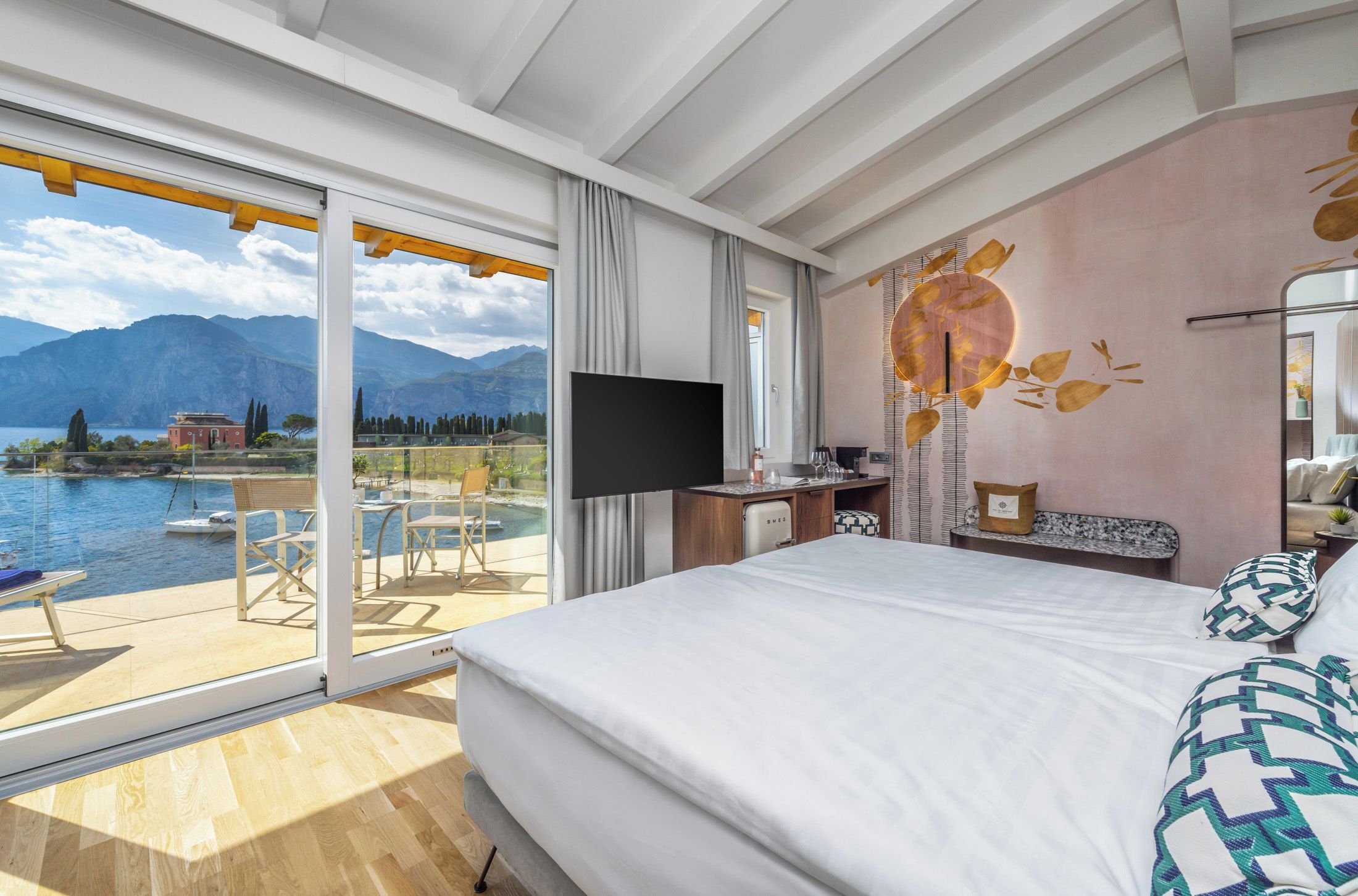 Habitación de hotel con una cama grande, un televisor de pantalla plana, un escritorio con computadora y vistas a un lago y montañas.