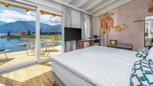 A hotel room with a large bed, a flat-screen TV, a desk with a computer, and a view of a lake and mountains. - Hotel Val di Sogno (Malcesine)