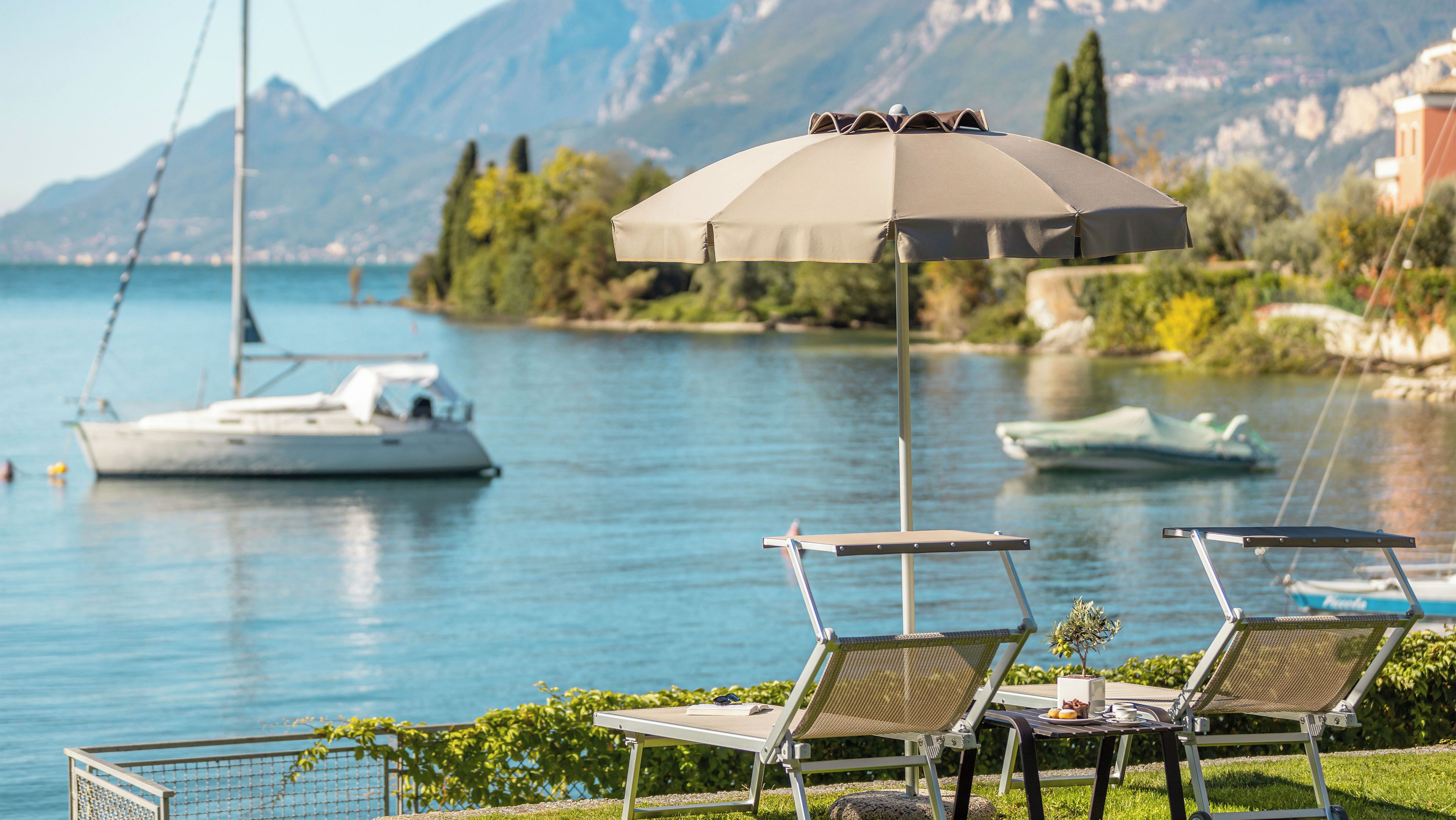 A lakeside view with two lounge chairs, an umbrella, and a table with a plant and a book.
