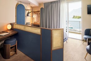 A hotel room with a desk, a chair, a small stool, a bed, a wardrobe, and a view of a lake through a balcony. - Hotel Val di Sogno (Malcesine)