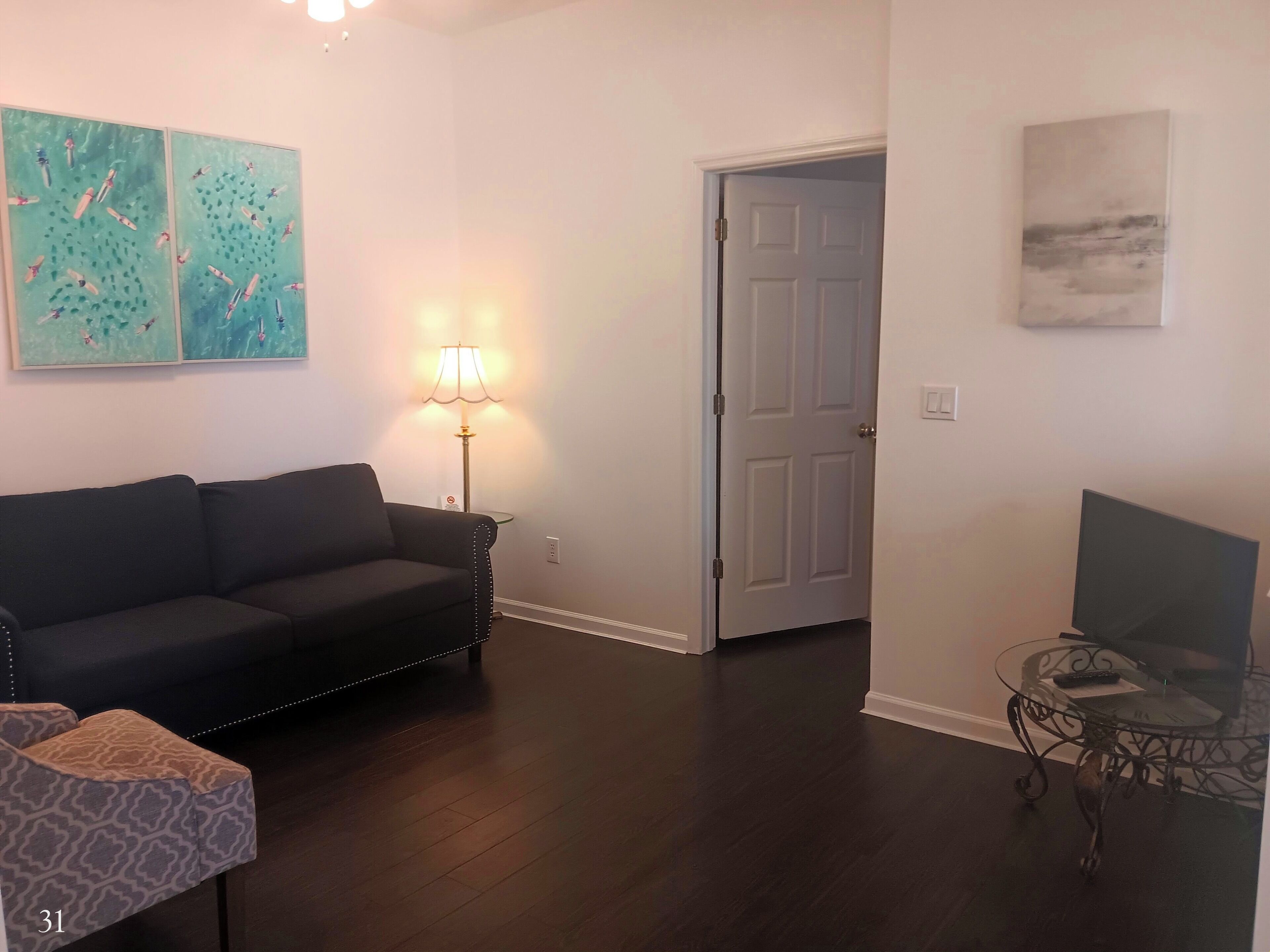 A living room with a dark wood floor, a black sofa, a patterned ottoman, a glass coffee table, a lamp, and two wall art pieces.