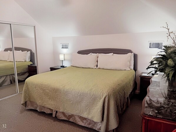 A bedroom with a large bed, a bedside table with a lamp, a mirror, and a potted plant.