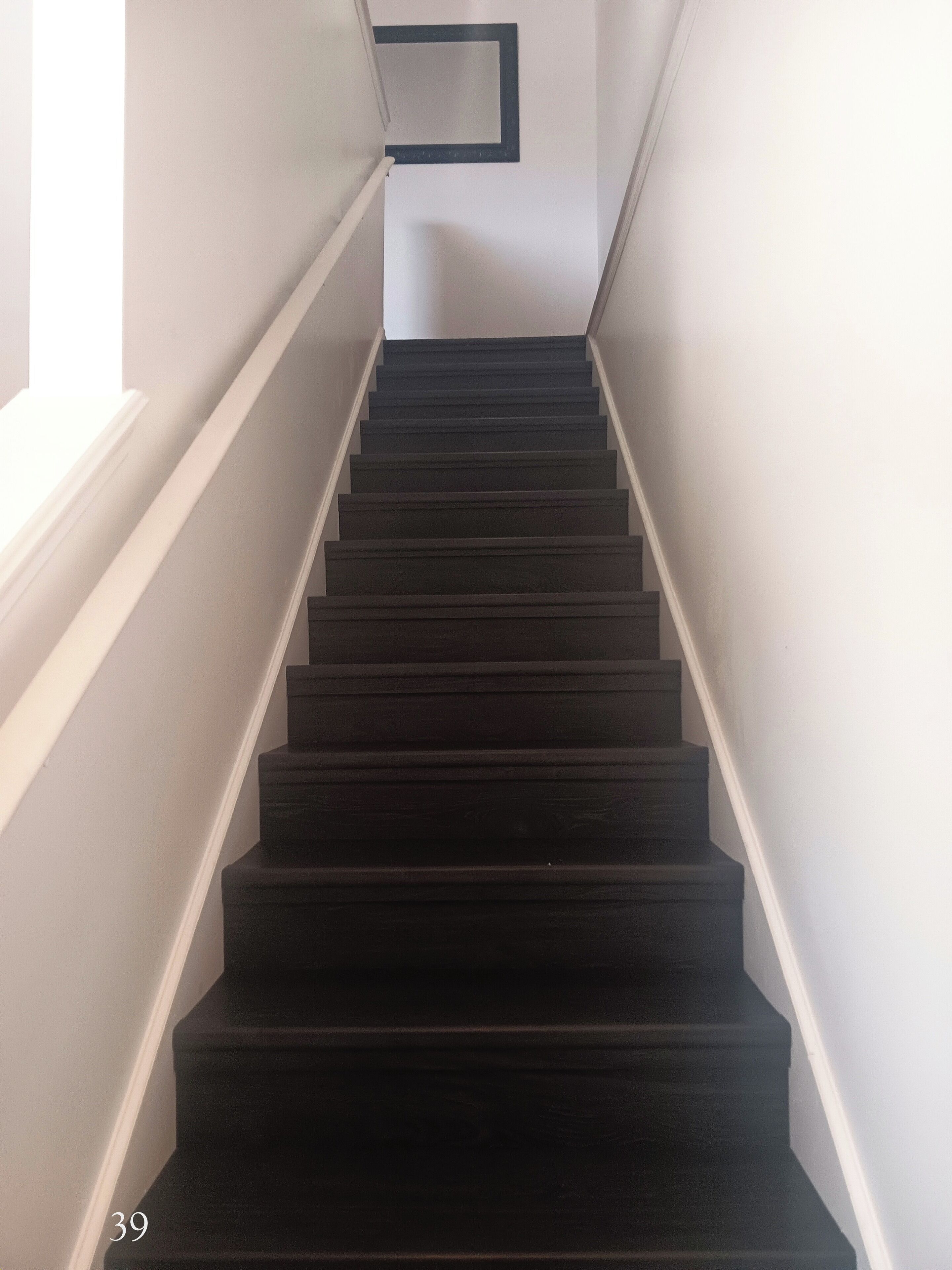 Uma escadaria com degraus escuros e corrimãos brancos, que leva a um patamar com um espelho emoldurado a preto.