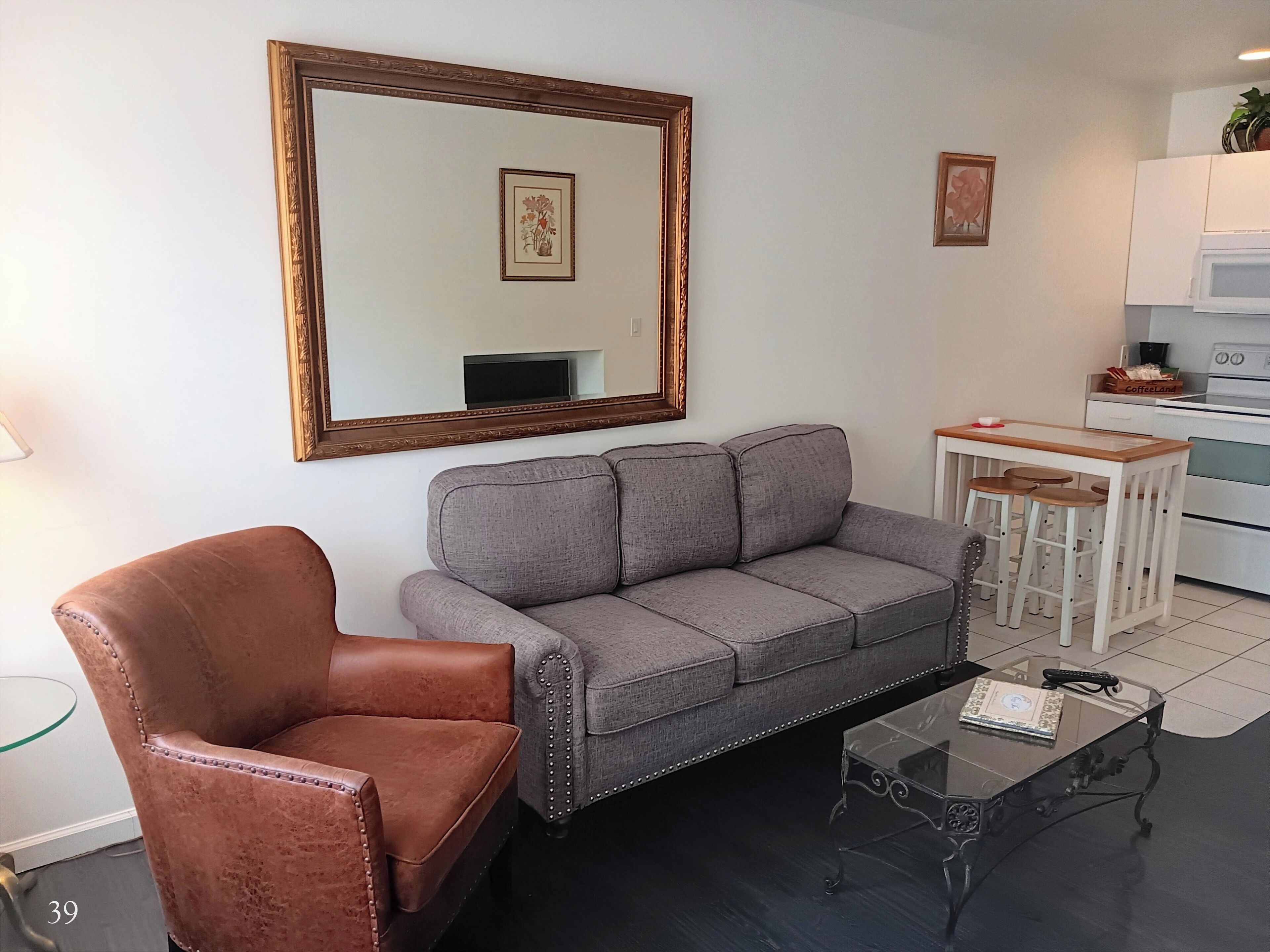 Una sala de estar con un sofá gris, un sillón de cuero marrón, una mesa de centro de vidrio, una zona de cocina con microondas y cafetera, y un espejo grande en la pared.