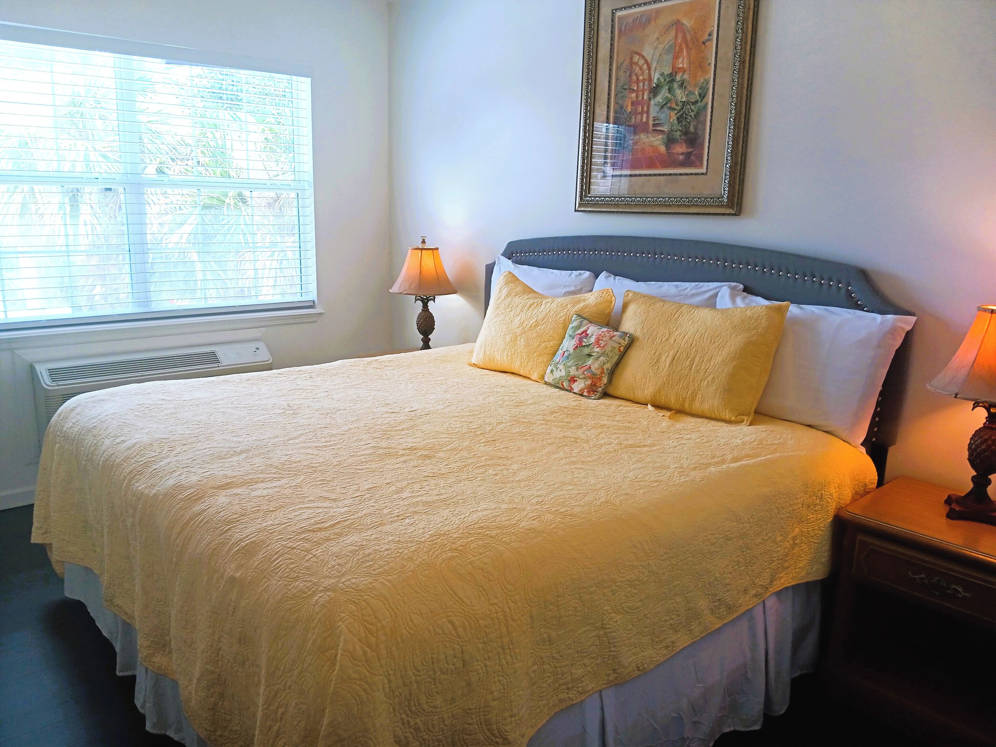 A neatly made bed with a patterned bedspread, a decorative pillow, and a headboard. There is a window with blinds, a bedside table with a lamp, and a framed picture on the wall.