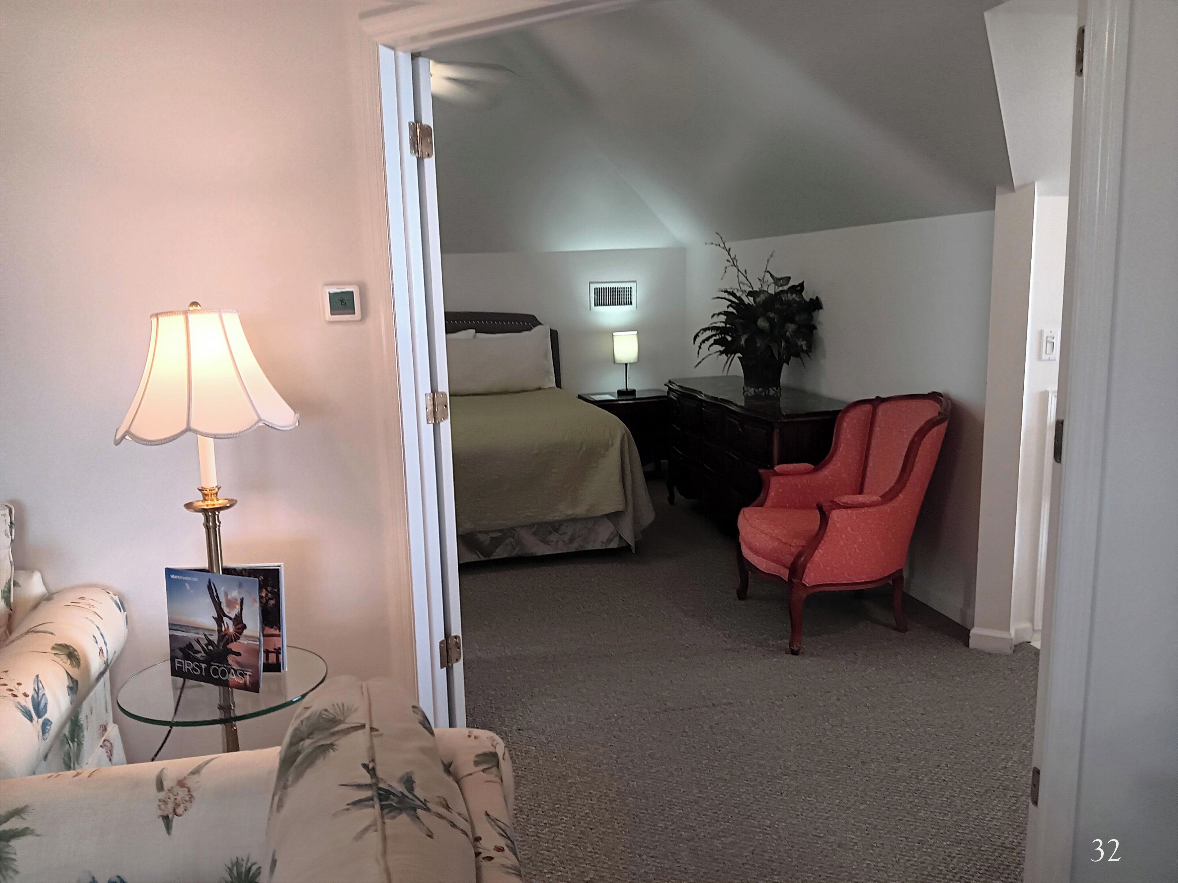 A hotel room with a bed, a chair, a lamp, a plant, and a framed picture on a table.