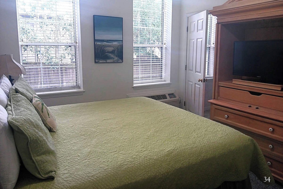 A bedroom with a bed, a television, and two windows with blinds.