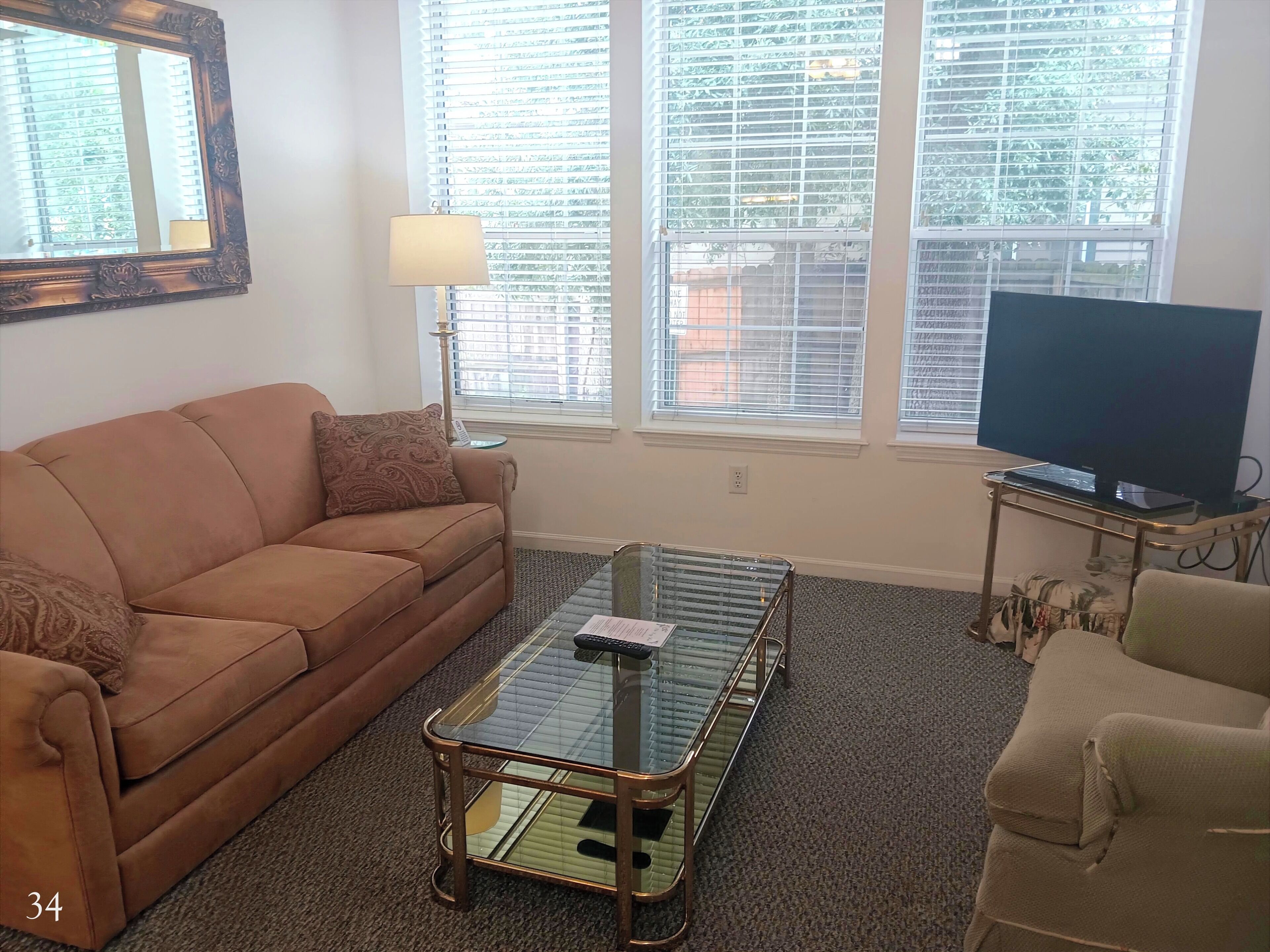 A living room with a brown sofa, a glass coffee table, a television, and a window with blinds.