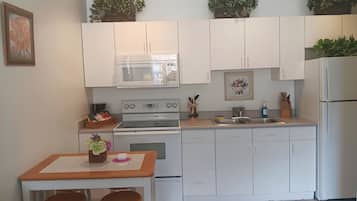 A compact kitchen with white cabinets, a microwave, an oven, a sink, and a refrigerator. There are two stools at a small table with a vase of flowers.