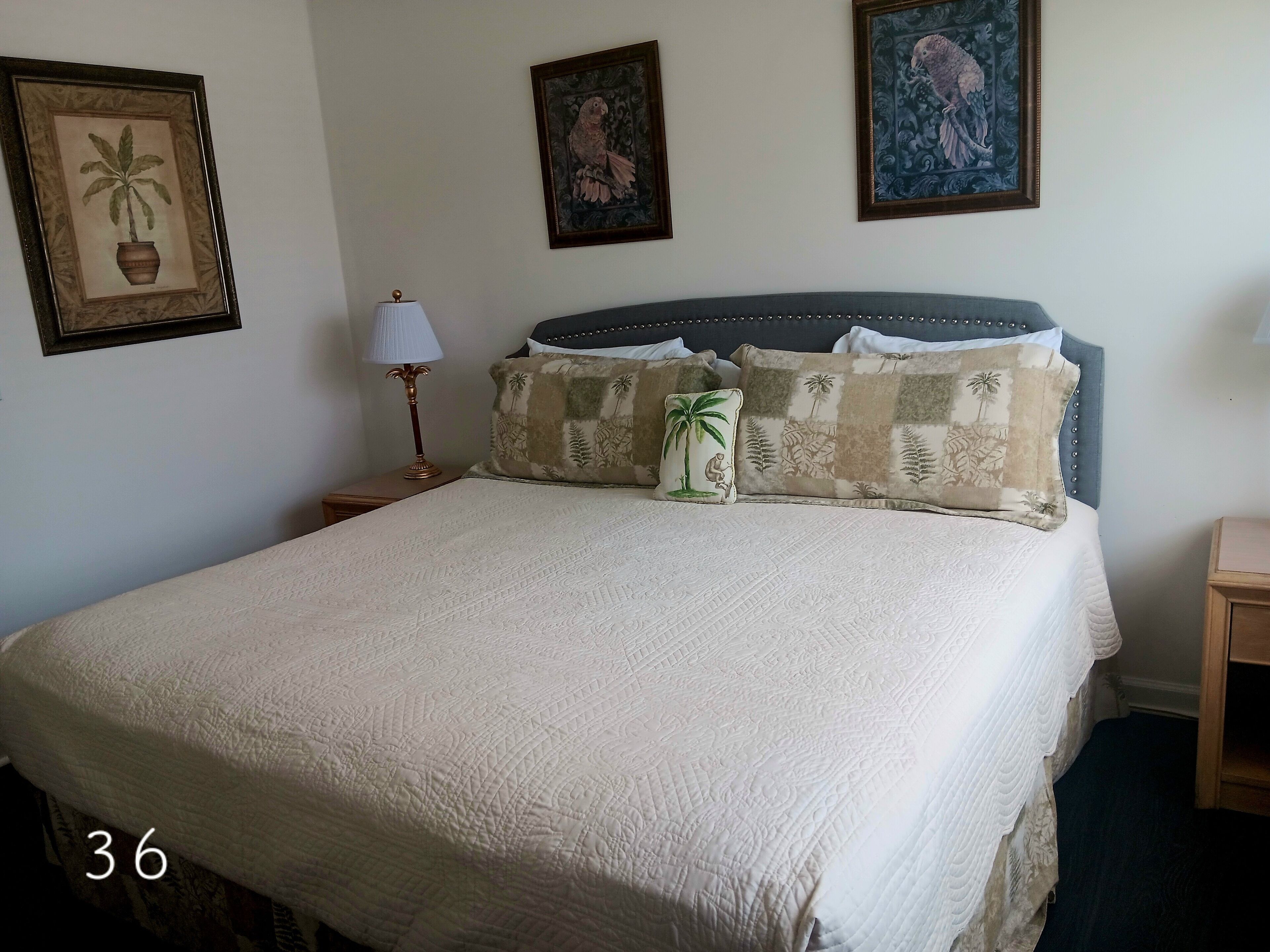 Una cama bien hecha con cubrecolcha blanco acolchado, un cojín decorativo con diseño de palmera y otro a juego con motivos tropicales. En la pared hay dos cuadros, uno con un pájaro y otro con fondo floral. Una lámpara de mesa con base dorada está sobre la mesita de noche junto a la cama.