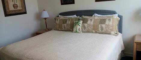 Una cama bien hecha con cubrecolcha blanco acolchado, un cojín decorativo con diseño de palmera y otro a juego con motivos tropicales. En la pared hay dos cuadros, uno con un pájaro y otro con fondo floral. Una lámpara de mesa con base dorada está sobre la mesita de noche junto a la cama.