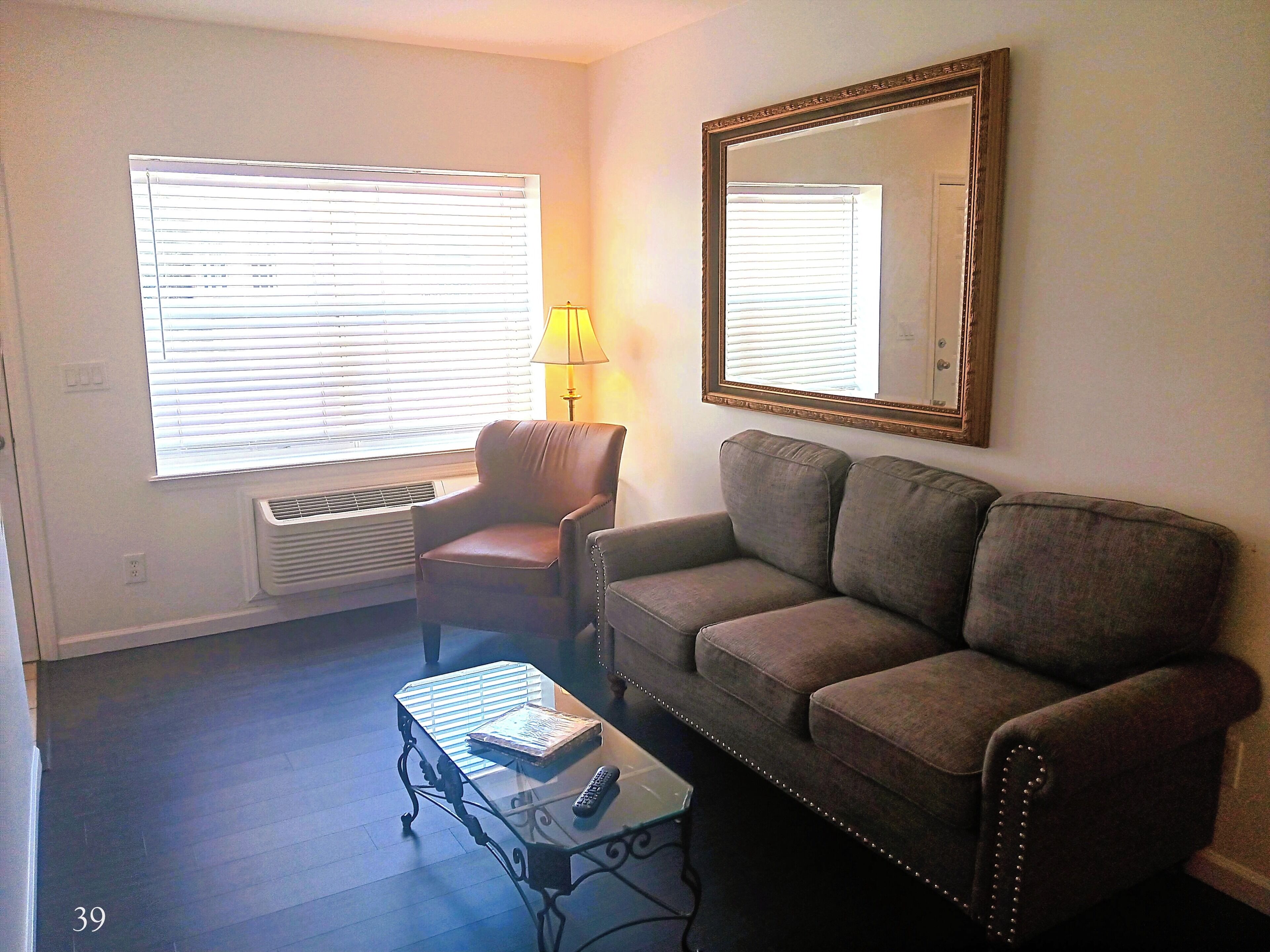 Un salon comprenant un fauteuil en cuir marron, un canapé gris, une table basse en verre avec un livre et une télécommande, une lampe et une grande fenêtre avec des stores.