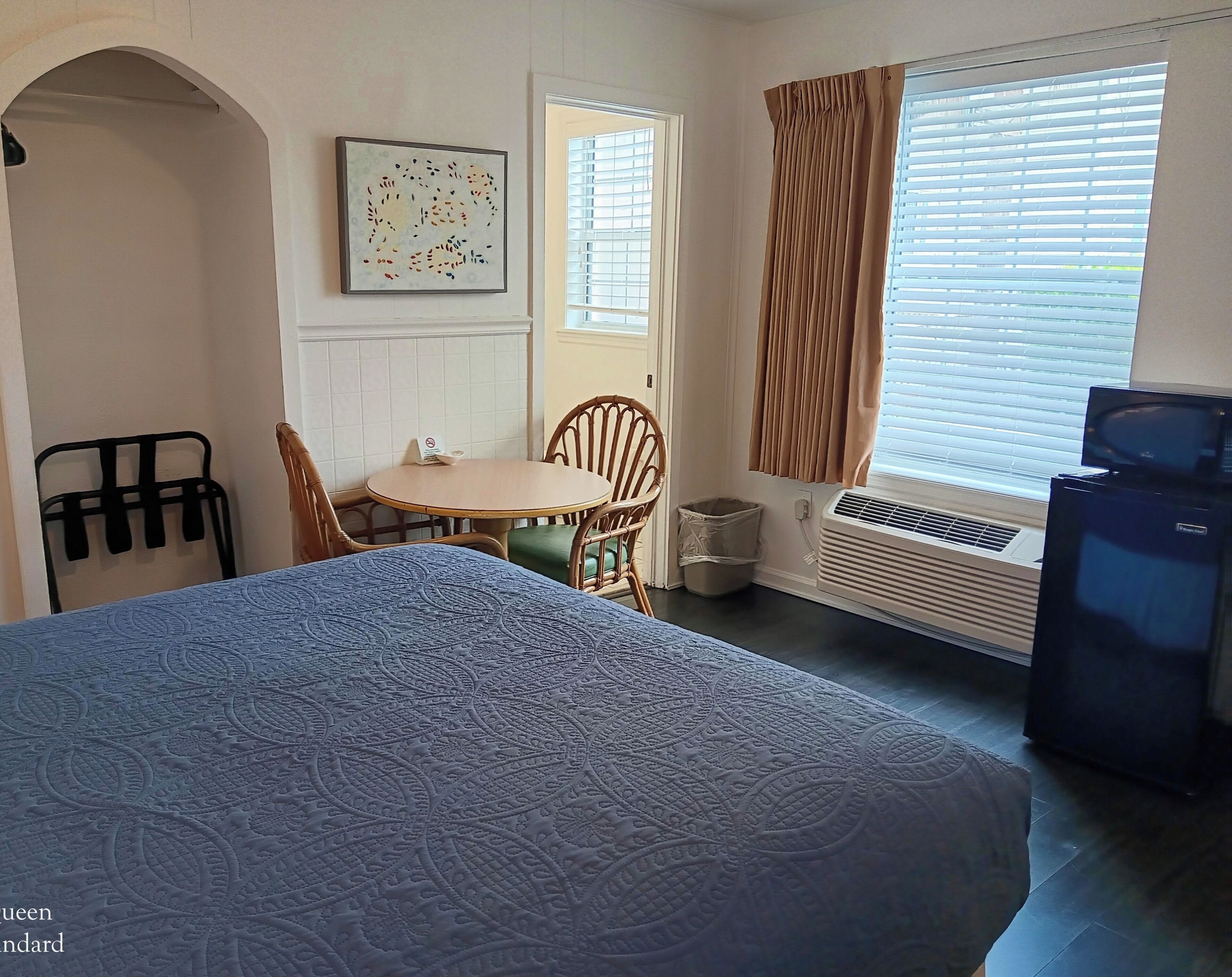 A room with a bed, a dining table with chairs, a window with curtains, an air conditioning unit, and a trash bin.