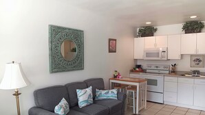 A compact living space with a grey sofa, a small dining table, a white kitchen with a microwave and oven, and a decorative mirror. - The Cozy Inn (St. Augustine)
