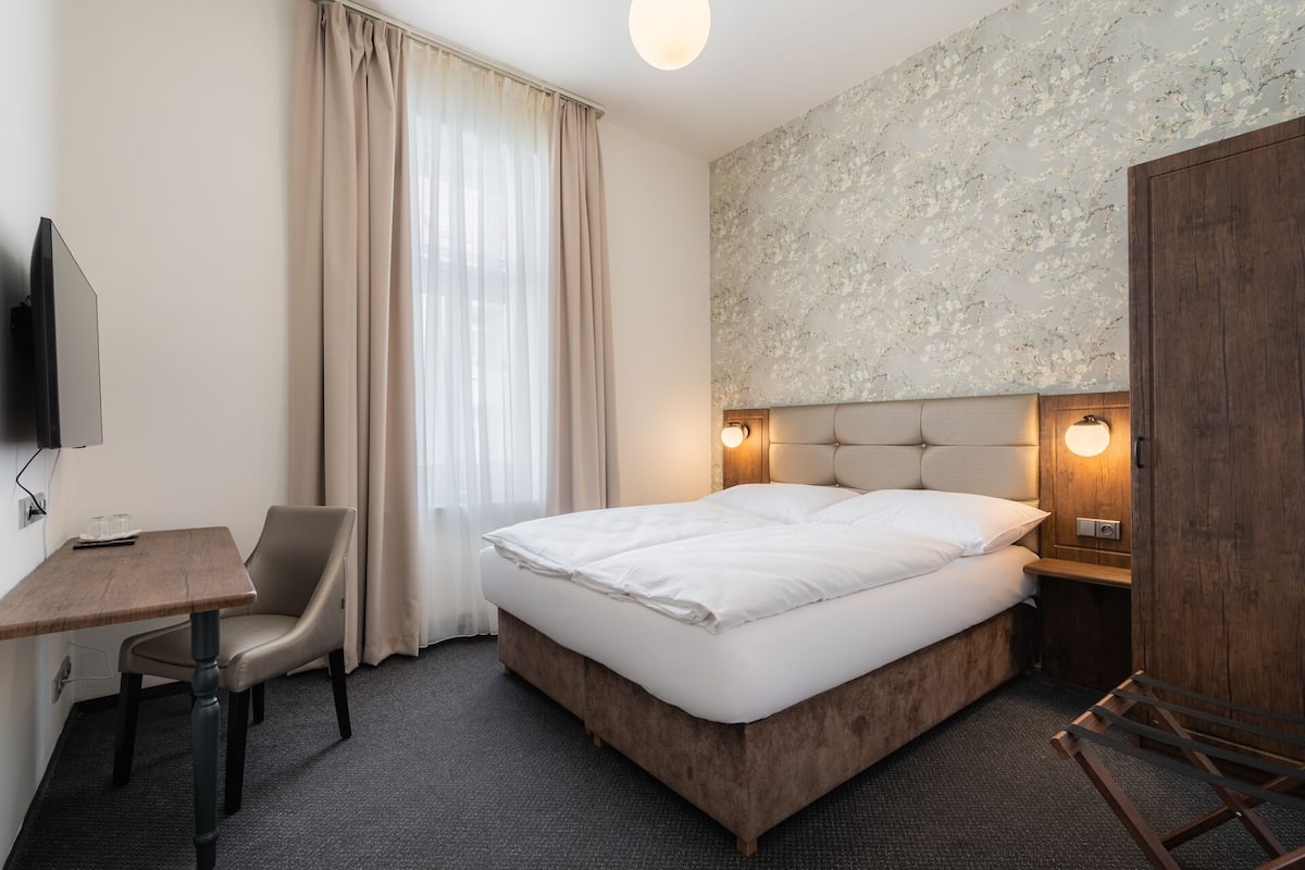 Quarto de hotel com cama, escrivaninha, cadeira, televisão e guarda-roupa.