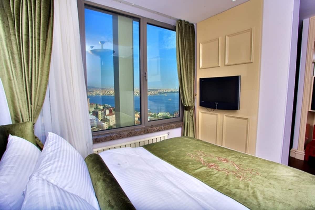 A hotel room with a large window, a bed with green bedding, a flat-screen TV mounted on the wall, and a view of a cityscape and water.