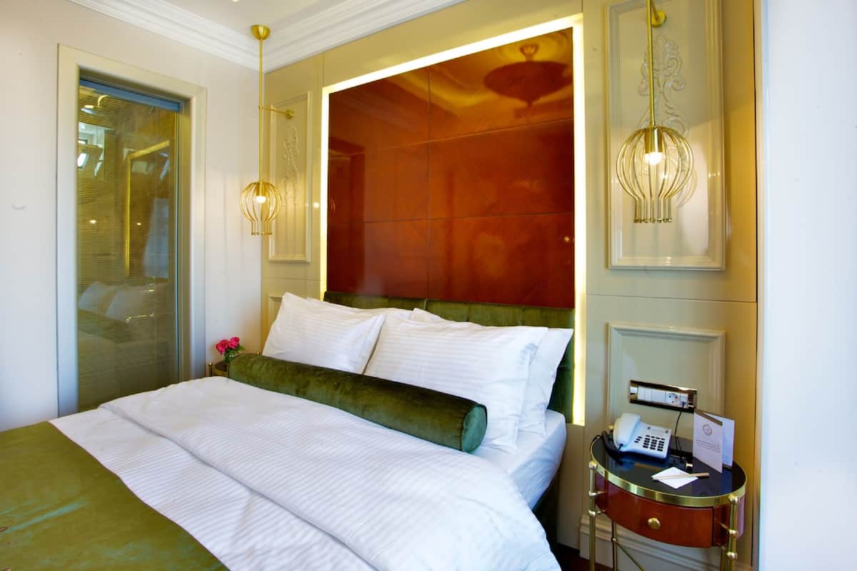 A hotel room with a large bed, a green accent pillow, a bedside table with a phone and papers, and a wall-mounted light fixture.
