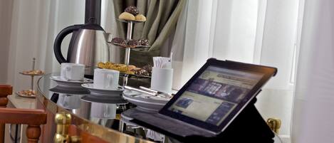 Colazione allestita con un laptop su un tavolo in vetro, una caffettiera e un vassoio per dolci.