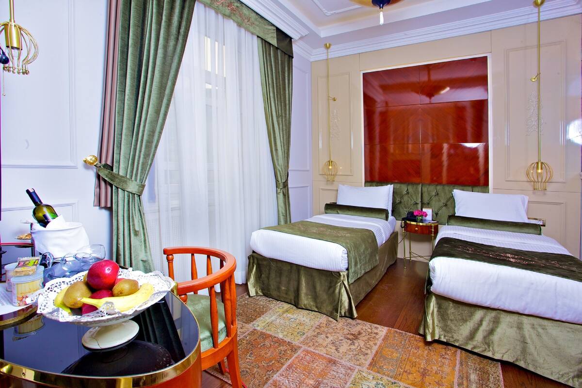 A hotel room with two beds, a dining table with fruit, a chair, and a small table with a bottle and glasses.