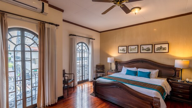 Um quarto espaçoso com uma cama grande, piso de madeira, ventilador de teto e varanda com grades de ferro forjado.