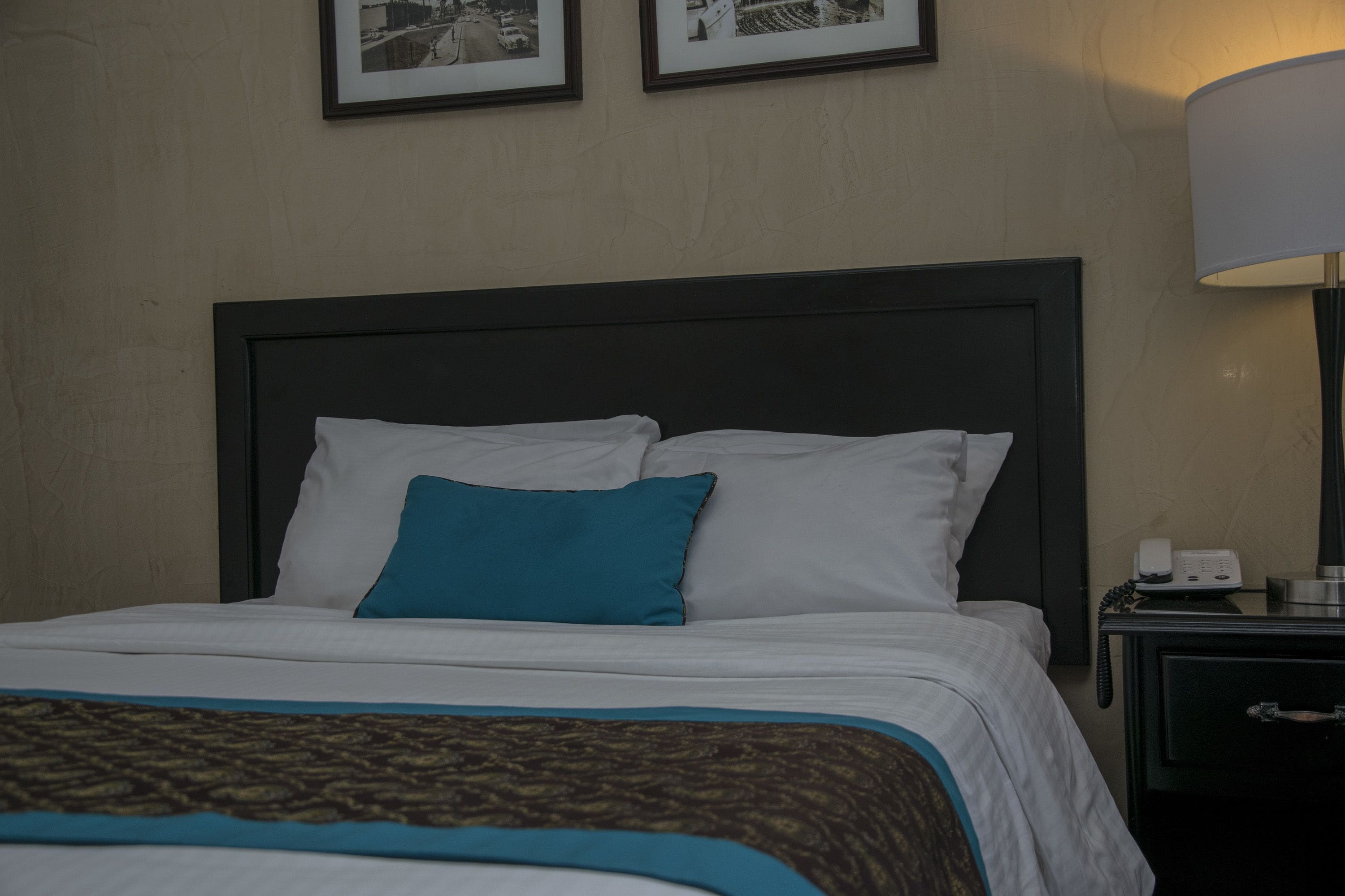 Een net opgemaakt bed met witte en teal kussens, een patroon geweven plaid en een nachtkastje met een lamp en telefoon.