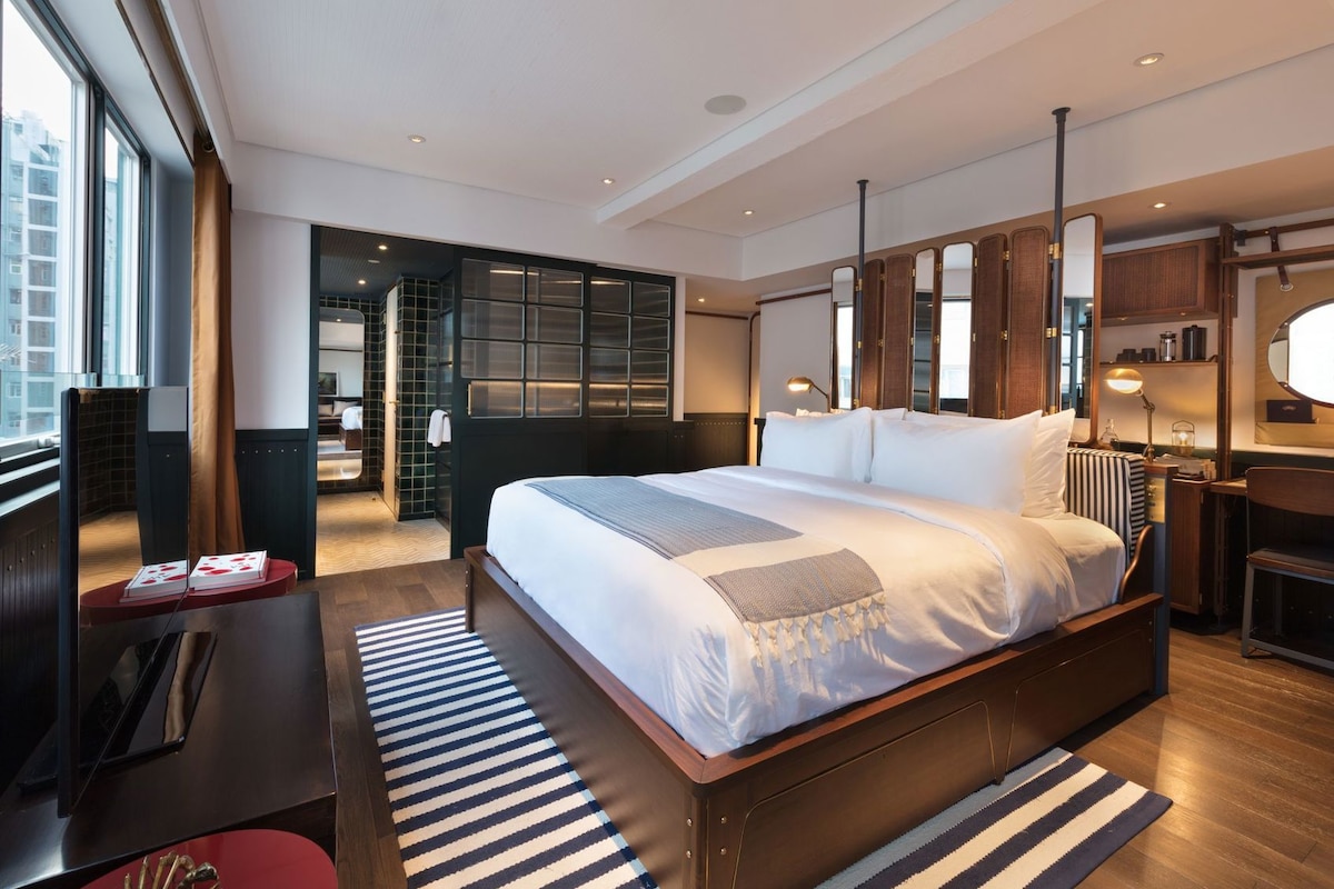 Habitación de hotel moderna con una cama grande, un escritorio, una silla, una mesita de noche, un espejo y vistas a la ciudad.