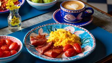 A breakfast setting with a plate of eggs, bacon, and tomatoes, a bowl of avocado, and a cup of coffee on a blue table.