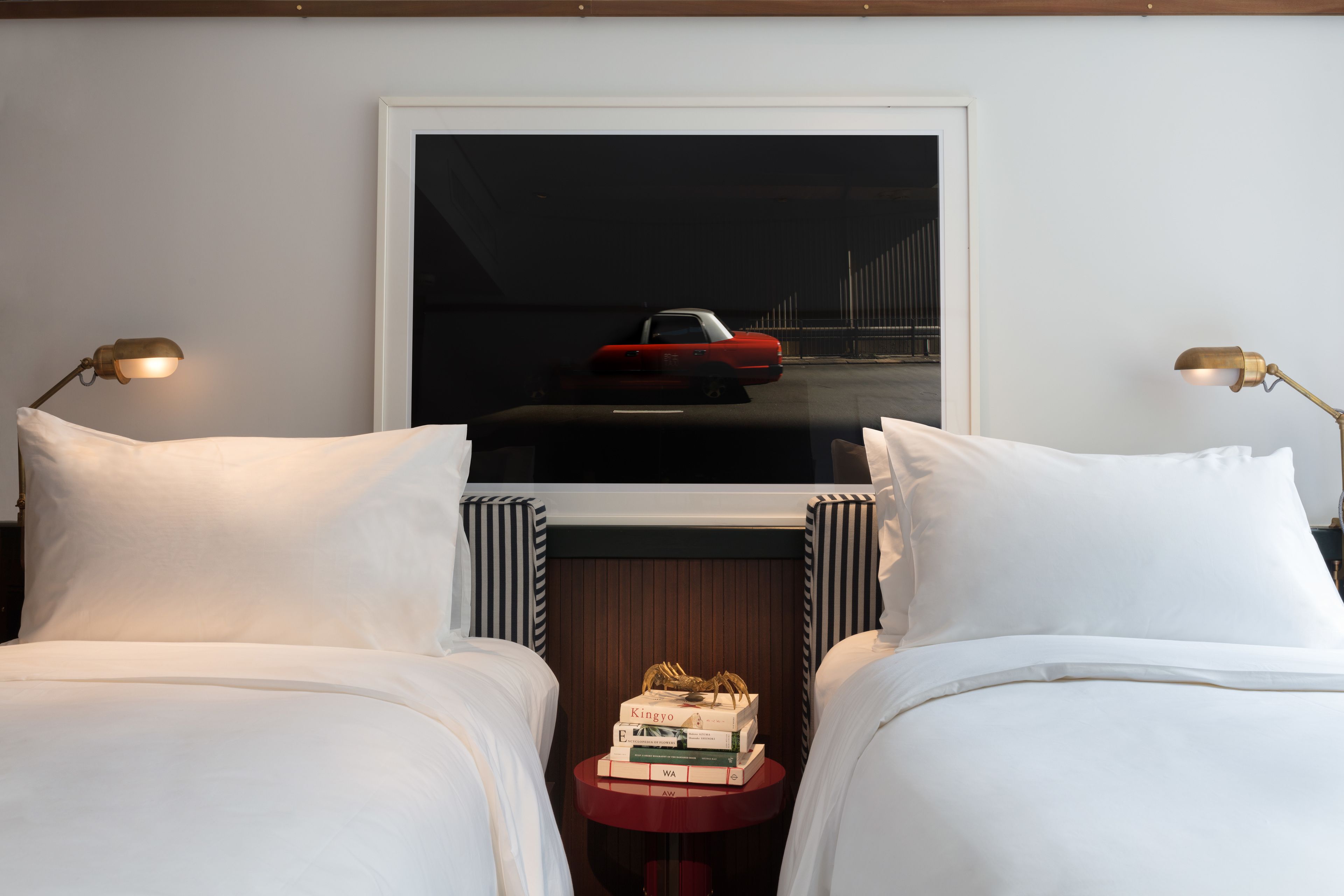 Een hotelkamer met twee bedden, een klein tafeltje met boeken en een ingelijst beeld van een rode auto op een brug.
