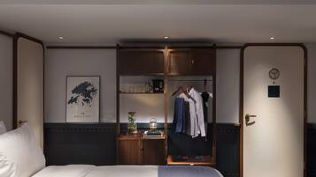 A hotel room with a bed, a wardrobe, a small table with a lamp, and a wall-mounted clock.