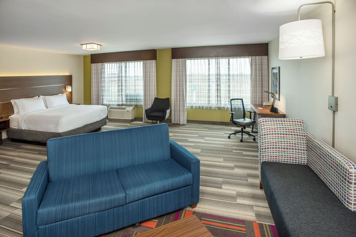 Une chambre d’hôtel avec un lit, un canapé bleu, un bureau avec une chaise et de grandes fenêtres avec des rideaux.
