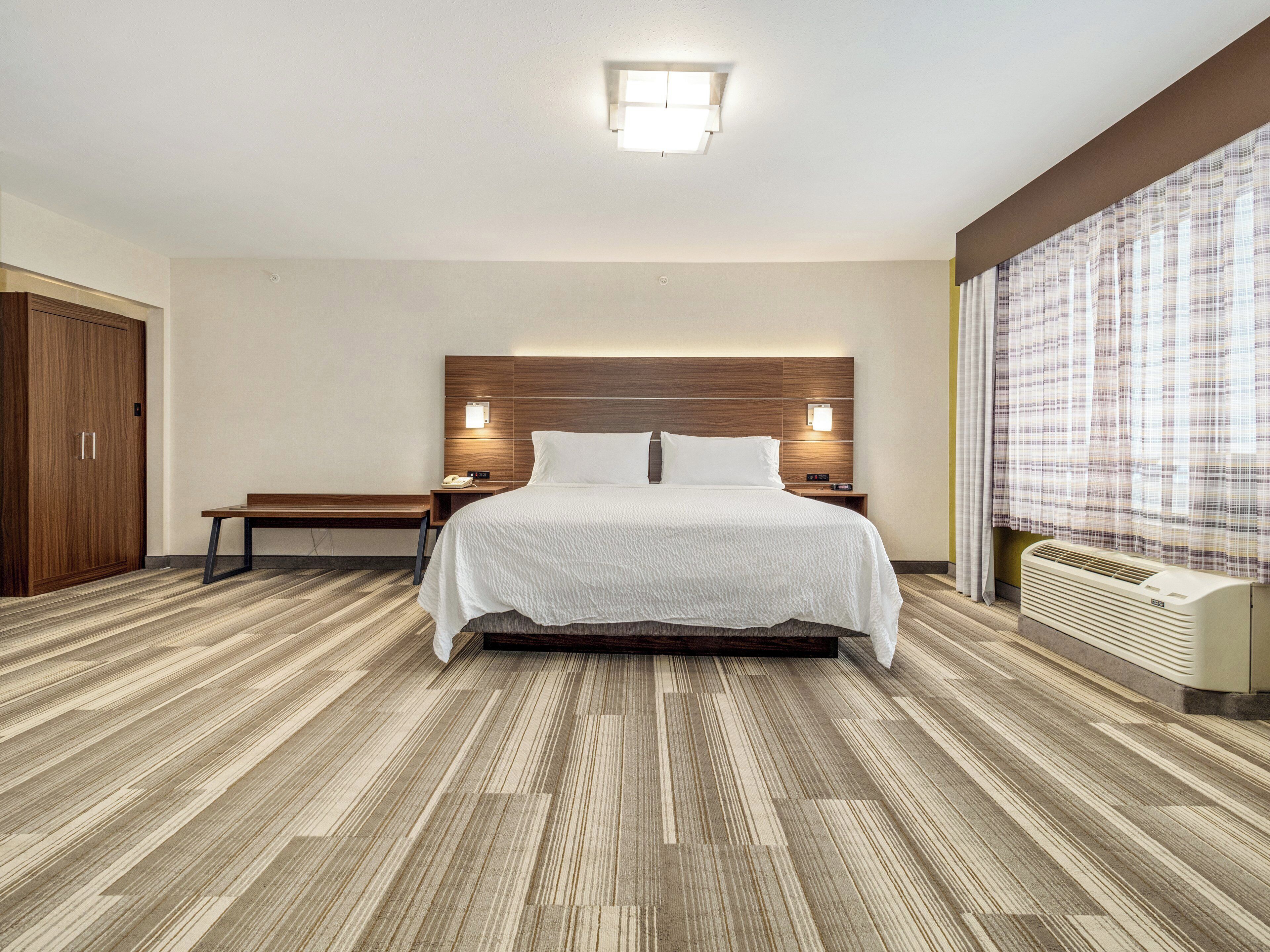Een hotelkamer met een groot bed, een houten hoofdeinde, nachtkastjes met lampen, een bankje, een garderobe en een groot raam met gordijnen.