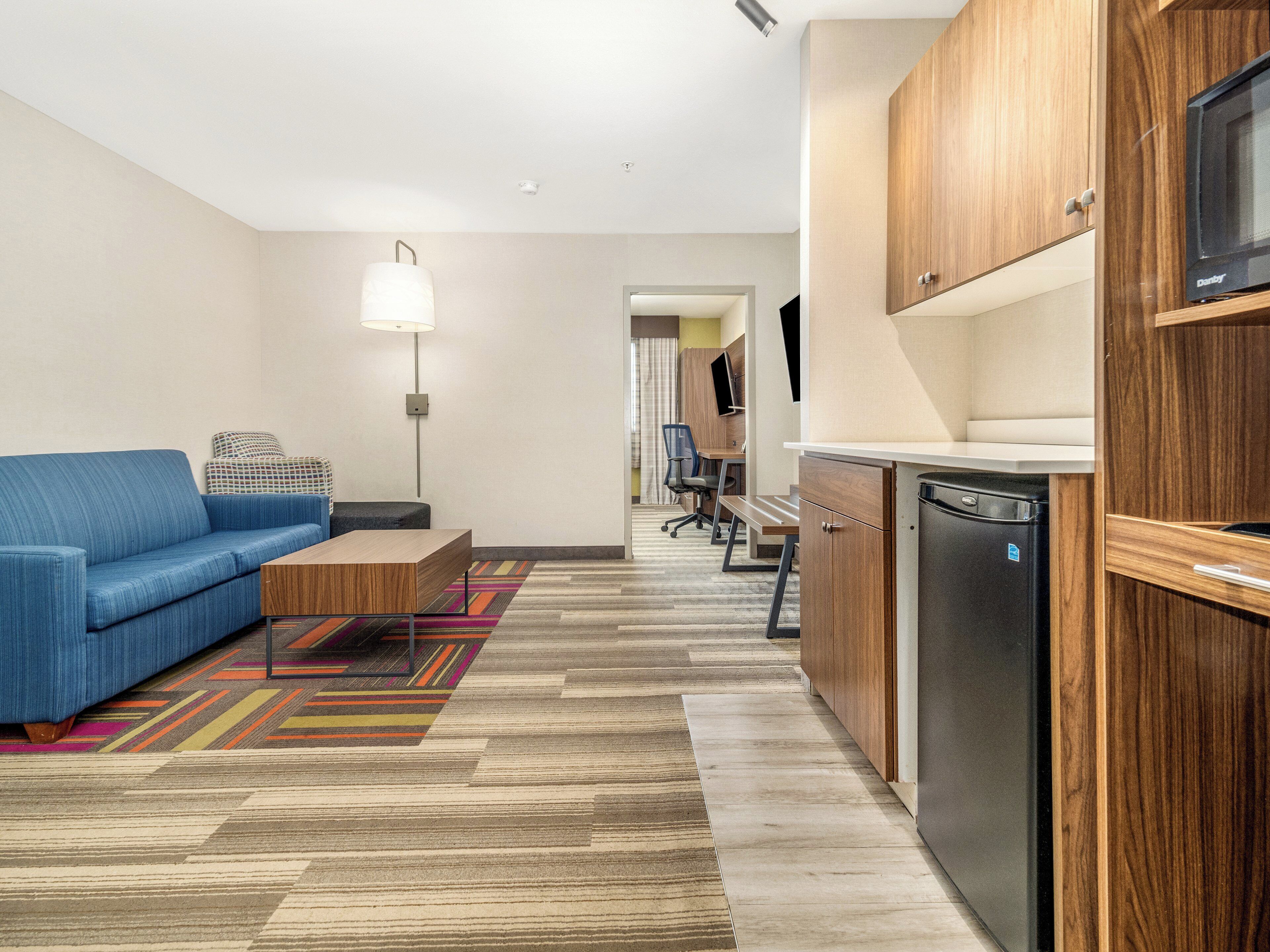 A modern hotel room with a blue sofa, a wooden coffee table, a colorful rug, a kitchenette with a microwave, and a desk with a chair.
