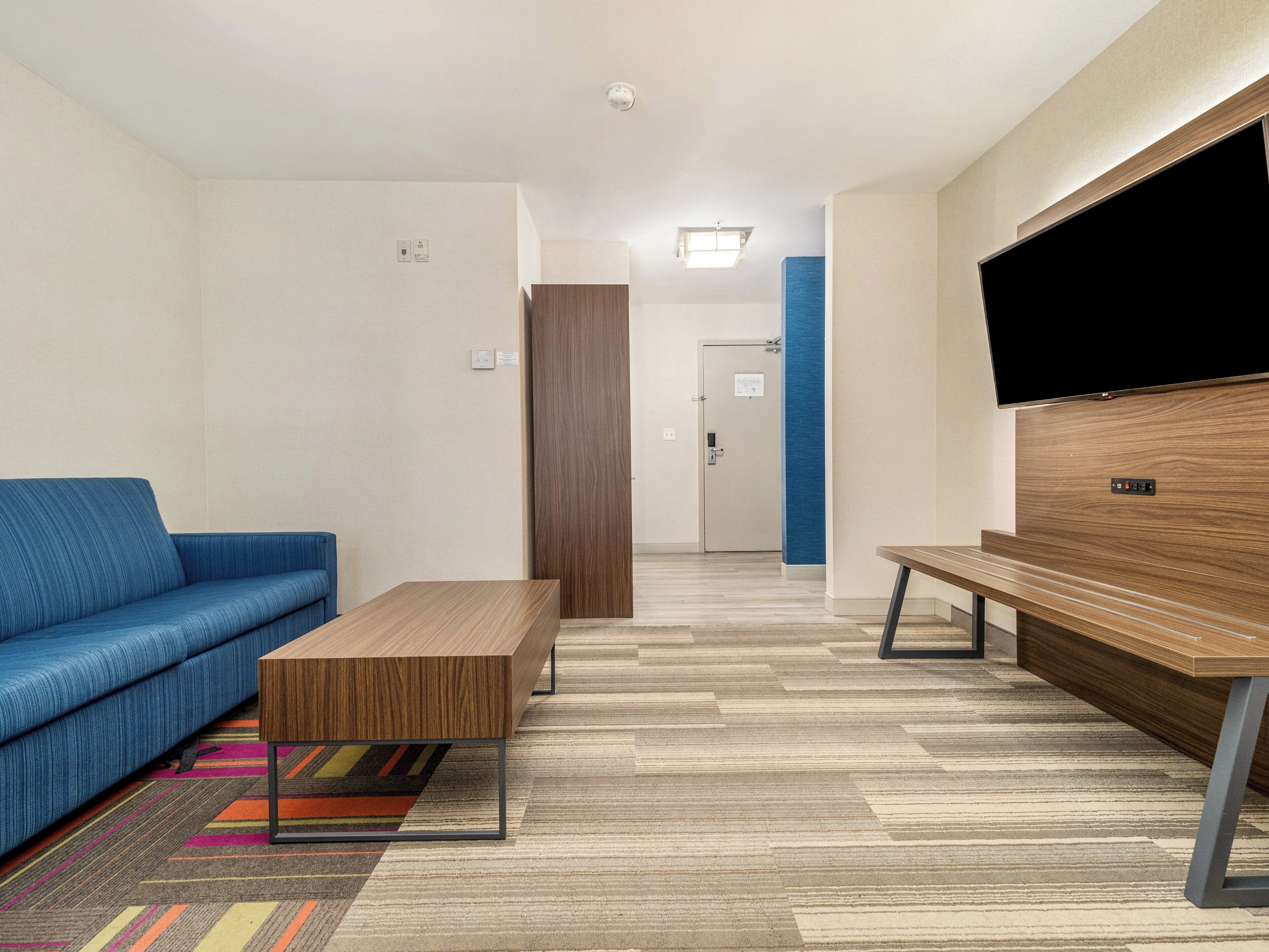 A modern hotel room with a blue sofa, a wooden coffee table, a bench, and a flat-screen TV mounted on a wooden wall.