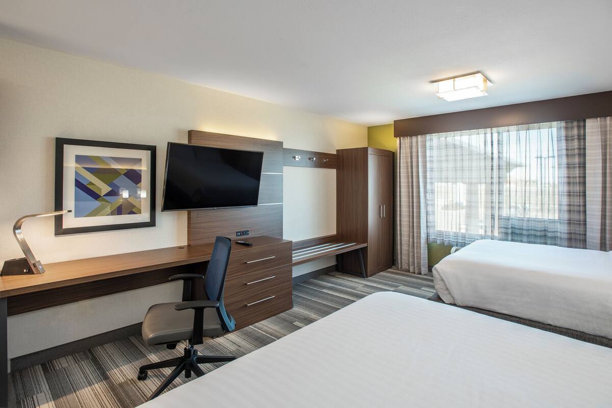 Une chambre d’hôtel comprenant un lit, un bureau avec une chaise, une télévision, une armoire et une fenêtre avec des rideaux.