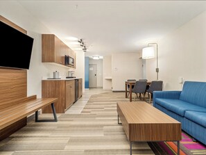 Een moderne hotelkamer met een houten bankje, een blauwe bank, een eettafel met stoelen, een magnetron en een lamp.
