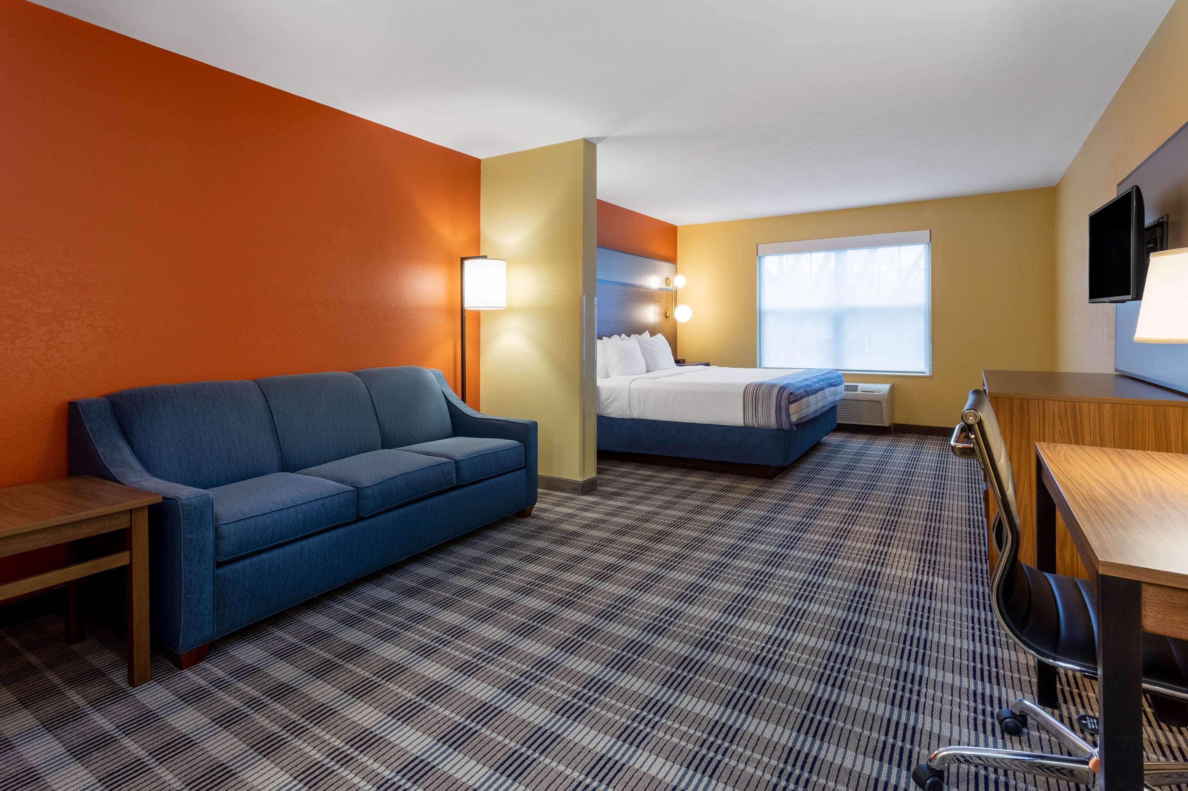 Habitación de hotel con sofá azul, cama con ropa de cama blanca, escritorio con silla y televisor en la pared.