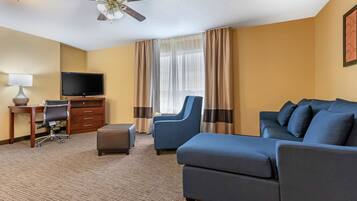 Habitación de hotel con un sofá grande, un escritorio con computadora, televisor y ventilador de techo.