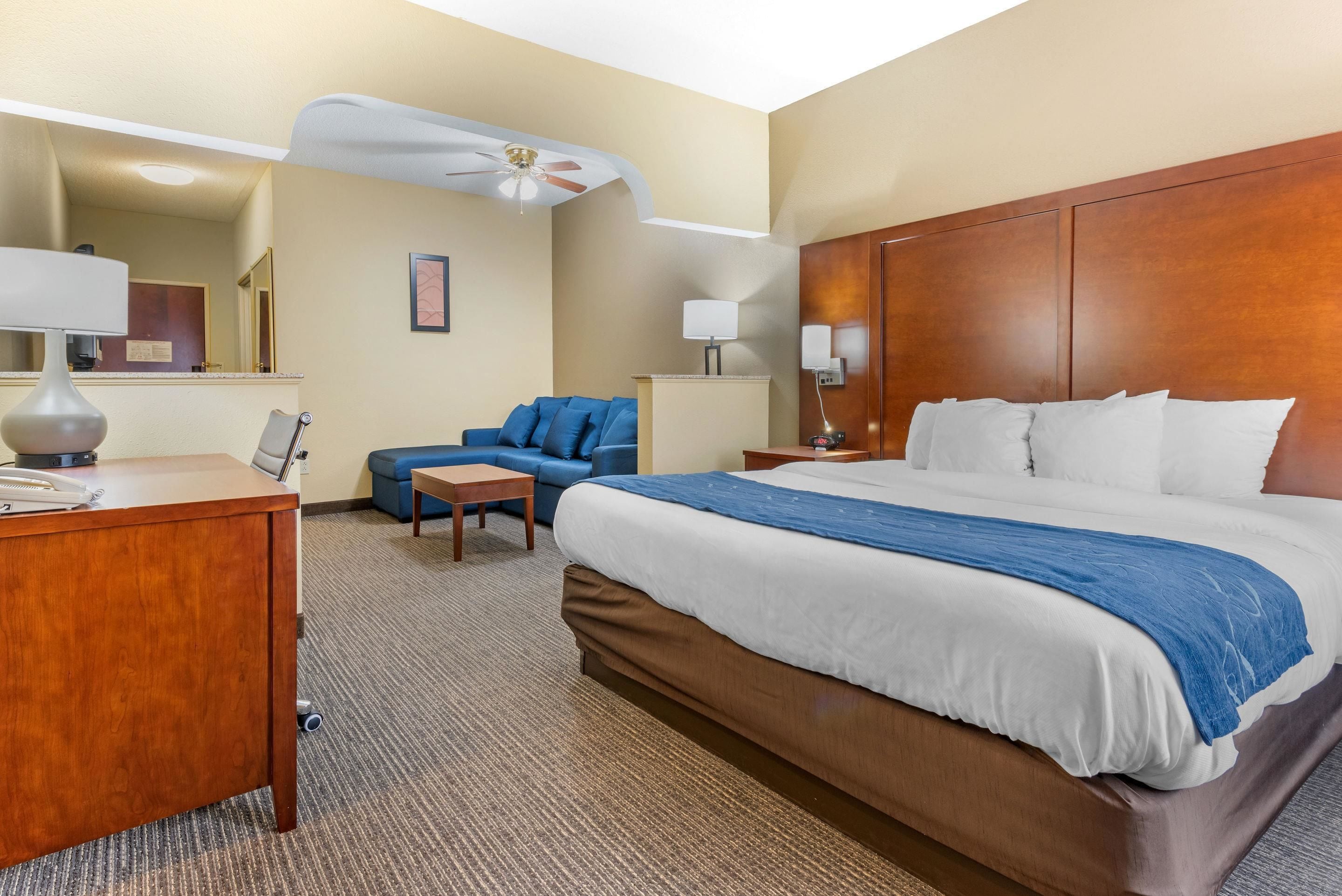 Une chambre d’hôtel avec un grand lit, un bureau avec une lampe, un canapé bleu, une petite table et un ventilateur de plafond.