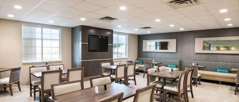 A spacious dining area with multiple tables and booths, a flat-screen TV mounted on the wall, and large windows providing natural light.