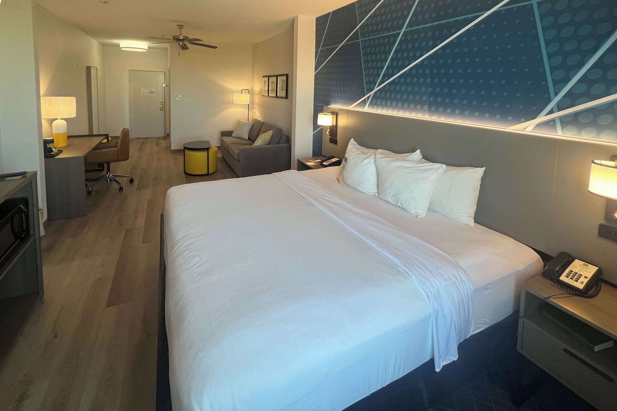 Ett hotellrum med en stor säng, ett skrivbord, en stol, ett litet bord, en lampa, en telefon och en vägg med ett geometriskt mönster.