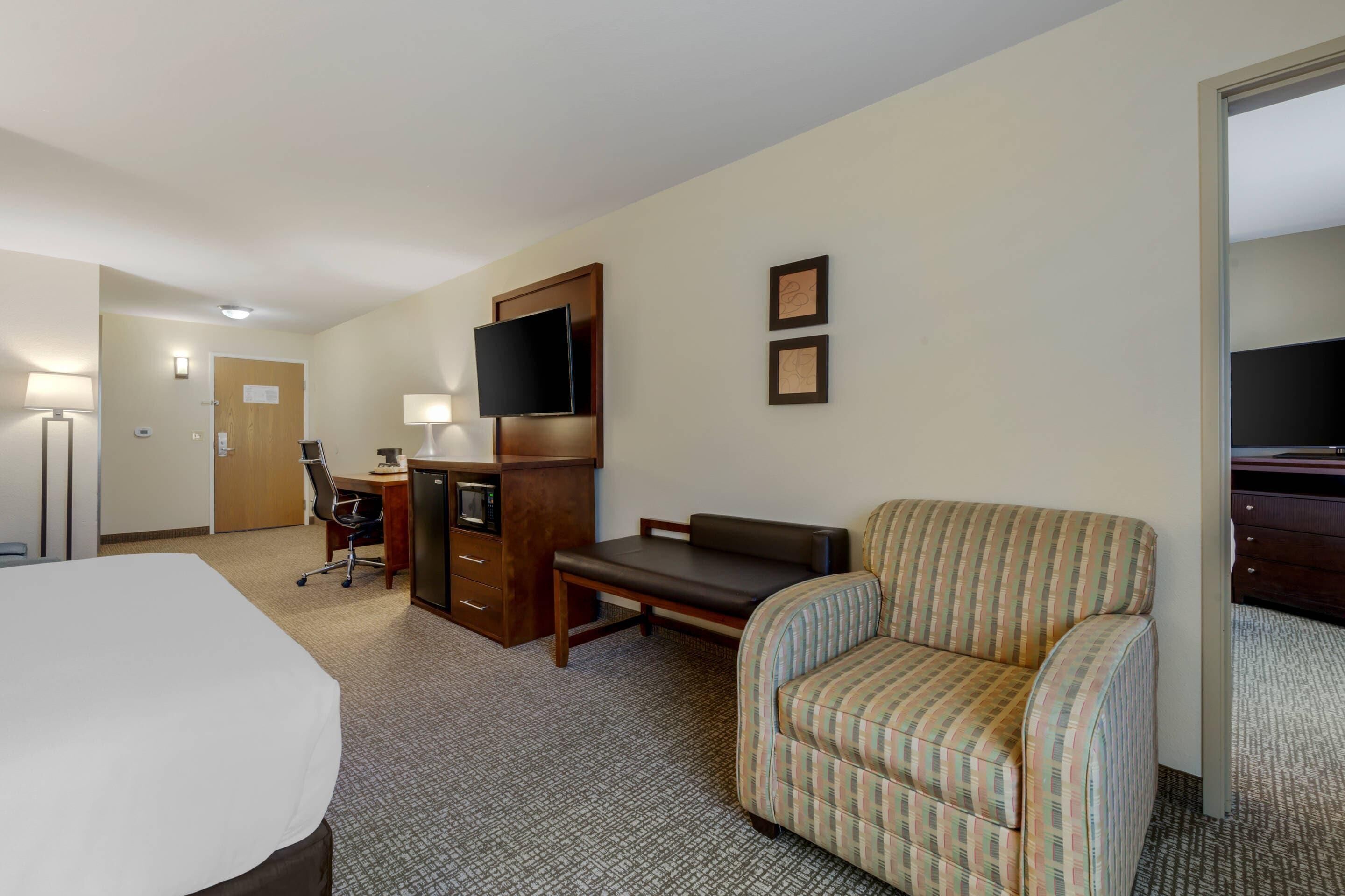 A hotel room with a bed, desk, chair, TV, and a small kitchenette.