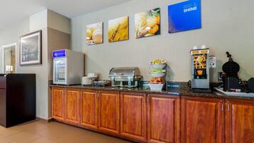 Una cocina de hotel con una estación de café, un fabricante de gofres y un bol de fruta.
